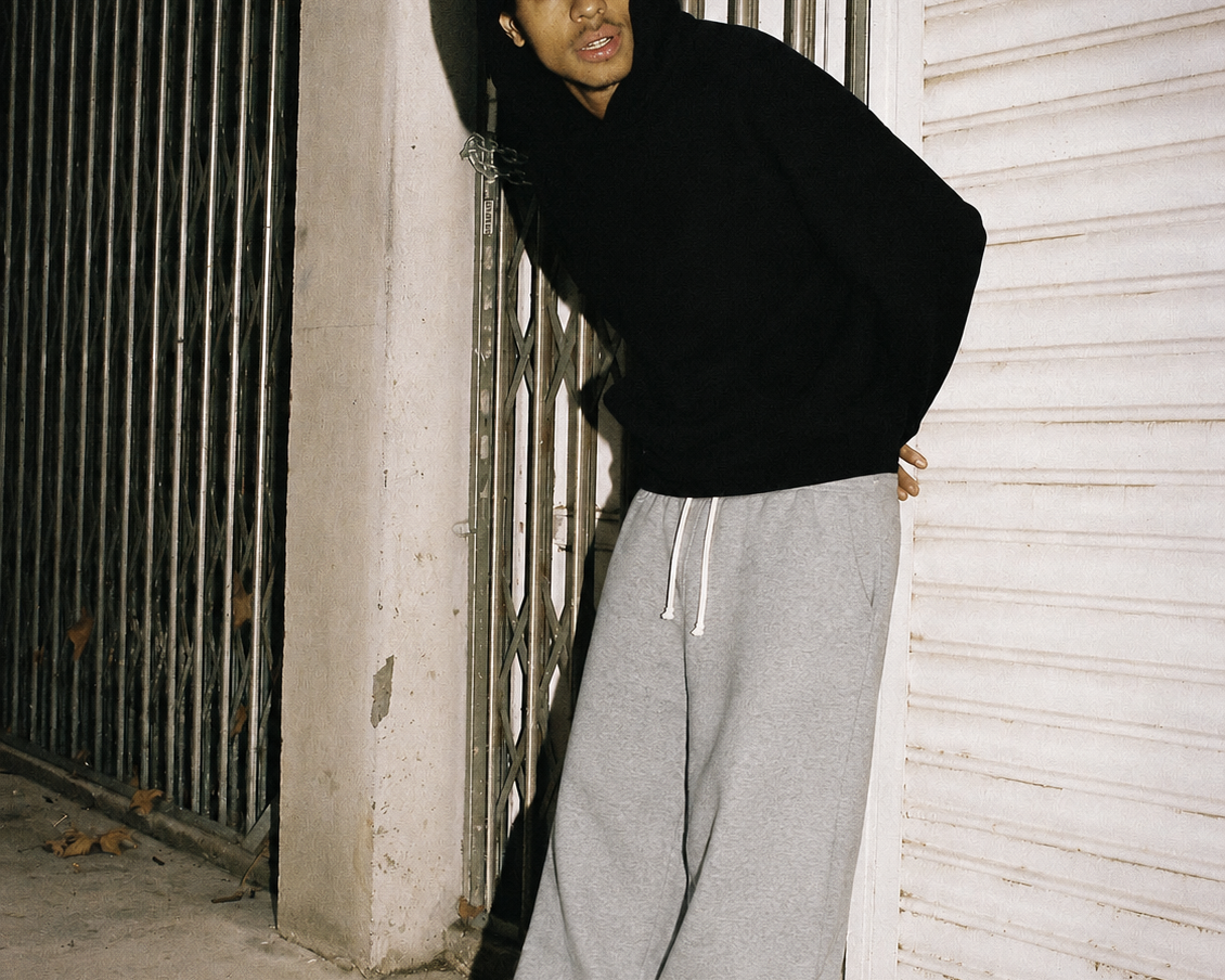 A person leaning against a metal gate and concrete wall at night, wearing a black hoodie, loose gray sweatpants, and white sneakers.